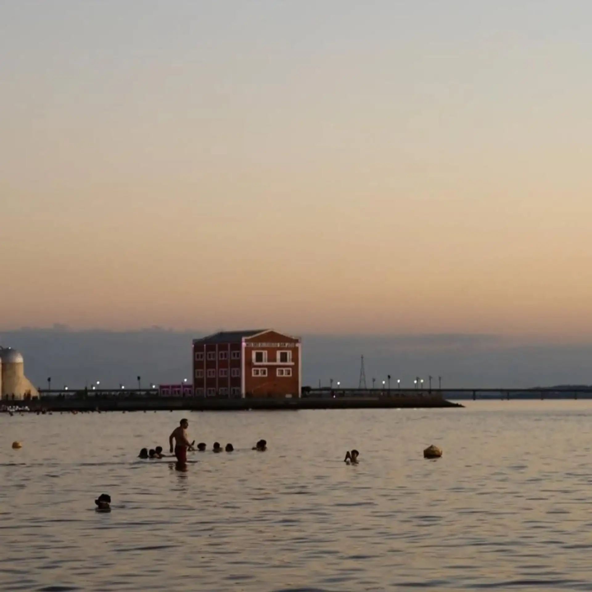 People swim in calm river water at dusk near a historic riverside building
