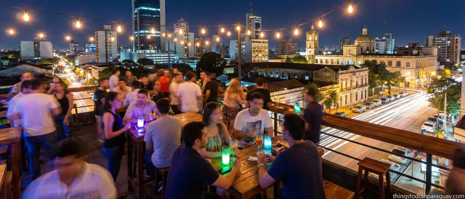 Crowded rooftop bar with string lights overlooking the illuminated Asuncion skyline at night