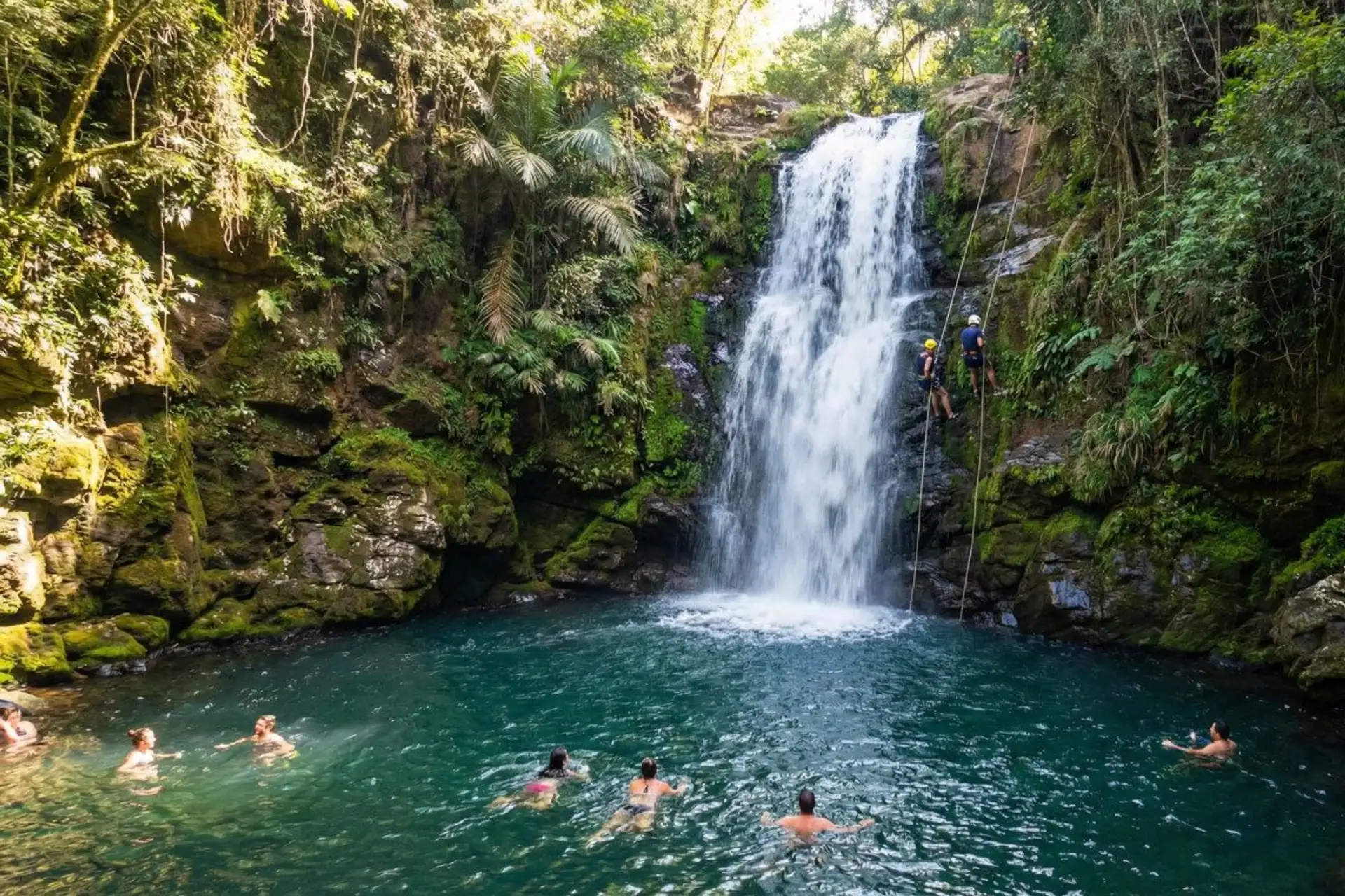 People swim in a turquoise pool beneath the Salto Cristal waterfall while another rappels down